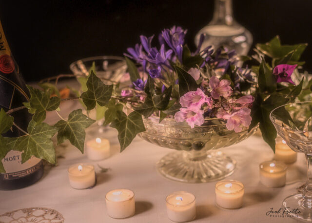 Photographie en gros plan d'une table illuminée par des bougies, avec une coupe de cristal remplie de fleurs violettes, du lierre et une bouteille de champagne.
