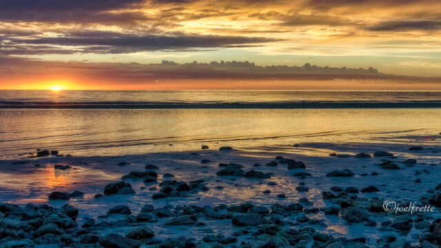 Photographie de paysage marin au coucher du soleil, avec des rochers sur le sable mouillé au premier plan et une lueur dorée flamboyante à l'horizon.