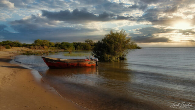 Photographie d'une barque rouge échouée sur une plage de sable au bord de l'eau, sous un ciel nuageux traversé par les rayons d'un soleil couchant