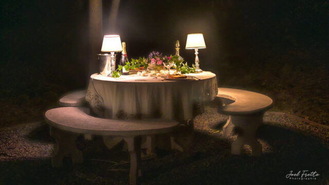 Photographie nocturne d'une table ronde élégamment dressée et éclairée par de petites lampes, entourée de bancs en pierre au milieu de l'obscurité totale.