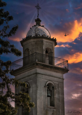 Photographie en contre-plongée d'un clocher d'église ancien au crépuscule, surmonté d'une croix avec des rayons lumineux, une cloche visible dans l'arcature et un oiseau volant dans le ciel orangé.