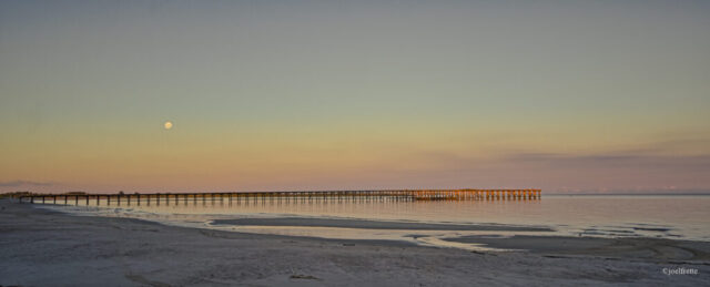 Photographie panoramique d'une longue jetée s'étendant sur l'eau calme au crépuscule, avec une lune brillante dans le ciel aux nuances douces et le sable de la plage au premier plan.