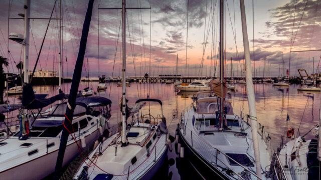 Photographie d'un port de plaisance rempli de voiliers au crépuscule, avec un ciel teinté de rose et de mauve se reflétant sur l'eau calme.