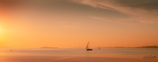 Photographie panoramique d'un voilier naviguant sur une mer calme au coucher du soleil dans une douce lumière aux tons orangés.