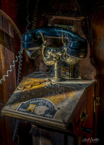 Photographie rapprochée d'un vieux téléphone mural en bois ancien envahi par des toiles d'araignée, avec une lumière rasante éclairant un autocollant commémoratif.