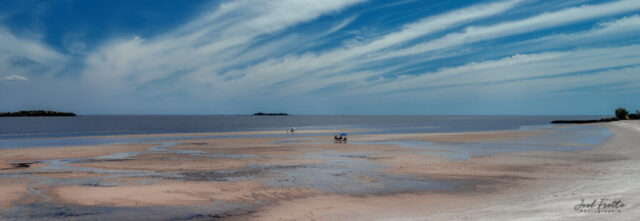 Photographie panoramique d'une grande plage de sable parsemée de reflets d'eau, sous un ciel bleu traversé de longs nuages filamenteux et un horizon lointain.