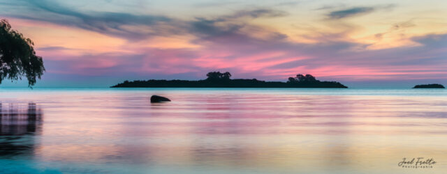 Paysage panoramique d'un plan d'eau calme avec des reflets de ciel rose et pastel au crépuscule et une petite île à l'horizon
